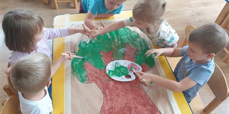 Powiększ grafikę: Dzieci siedzą przy stoliku i malują farbami drzewo