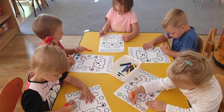 Powiększ grafikę: Dzieci z grupy II siedzą przy stoliku i kolorują kolorowanki z misiem i kropkami.