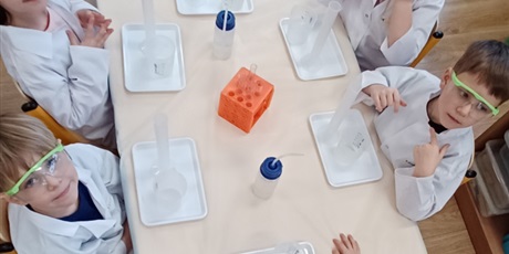 Powiększ grafikę: Zajęcia chemiczne w grupie IV - dzieci w białych fartuchach i okularach siedzą przy stolikach i przeprowadzają doświadczenia chemiczne