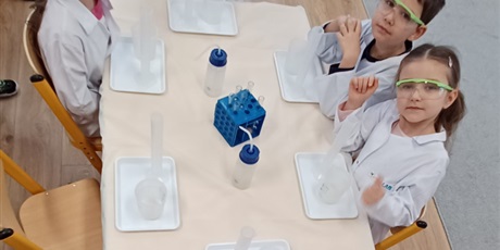 Powiększ grafikę: Zajęcia chemiczne w grupie IV - dzieci w białych fartuchach i okularach siedzą przy stolikach i przeprowadzają doświadczenia chemiczne