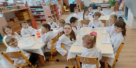 Powiększ grafikę: Zajęcia chemiczne w grupie IV - dzieci w białych fartuchach i okularach siedzą przy stolikach i przeprowadzają doświadczenia chemiczne