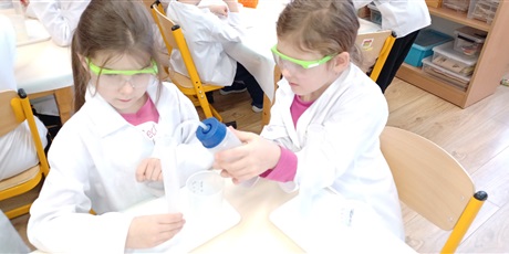 Powiększ grafikę: Zajęcia chemiczne w grupie IV - dzieci w białych fartuchach i okularach siedzą przy stolikach i przeprowadzają doświadczenia chemiczne