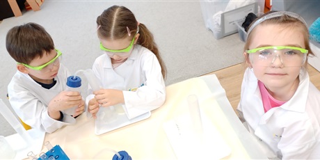 Powiększ grafikę: Zajęcia chemiczne w grupie IV - dzieci w białych fartuchach i okularach siedzą przy stolikach i przeprowadzają doświadczenia chemiczne