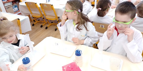 Powiększ grafikę: Zajęcia chemiczne w grupie IV - dzieci w białych fartuchach i okularach siedzą przy stolikach i przeprowadzają doświadczenia chemiczne