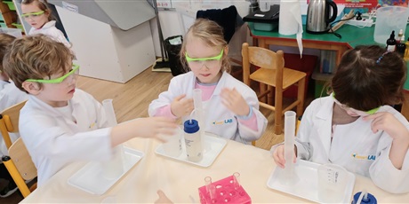 Powiększ grafikę: Zajęcia chemiczne w grupie IV - dzieci w białych fartuchach i okularach siedzą przy stolikach i przeprowadzają doświadczenia chemiczne