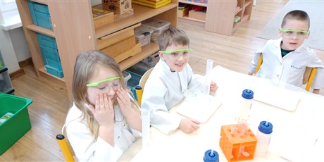 Powiększ grafikę: Zajęcia chemiczne w grupie IV - dzieci w białych fartuchach i okularach siedzą przy stolikach i przeprowadzają doświadczenia chemiczne