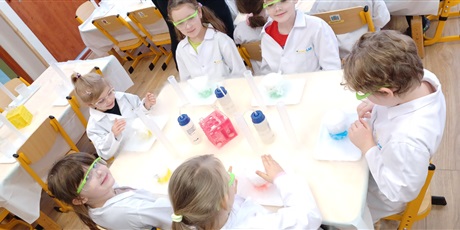 Powiększ grafikę: Zajęcia chemiczne w grupie IV - dzieci w białych fartuchach i okularach siedzą przy stolikach i przeprowadzają doświadczenia chemiczne