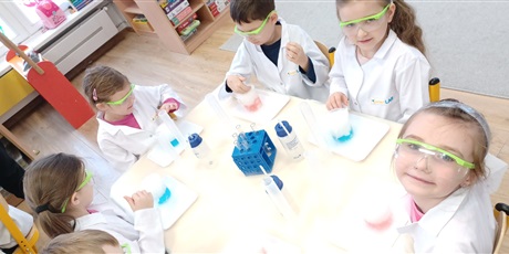 Powiększ grafikę: Zajęcia chemiczne w grupie IV - dzieci w białych fartuchach i okularach siedzą przy stolikach i przeprowadzają doświadczenia chemiczne
