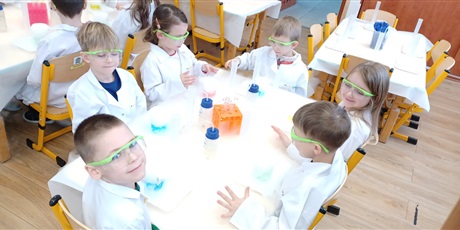 Powiększ grafikę: Zajęcia chemiczne w grupie IV - dzieci w białych fartuchach i okularach siedzą przy stolikach i przeprowadzają doświadczenia chemiczne