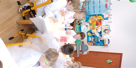 Powiększ grafikę: Zajęcia chemiczne w grupie IV - dzieci w białych fartuchach i okularach siedzą przy stolikach i przeprowadzają doświadczenia chemiczne