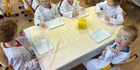 Powiększ grafikę: Zajęcia chemiczne w grupie III - dzieci w białych fartuchach i okularach siedzą przy stolikach i przeprowadzają doświadczenia chemiczne