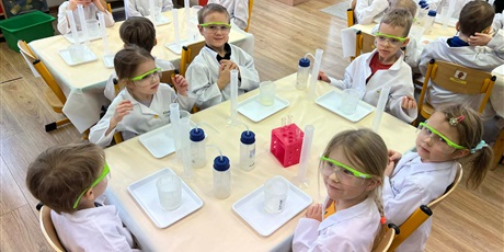 Powiększ grafikę: Zajęcia chemiczne w grupie III - dzieci w białych fartuchach i okularach siedzą przy stolikach i przeprowadzają doświadczenia chemiczne