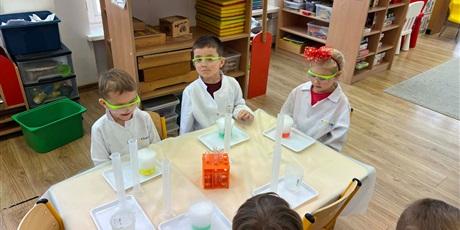 Powiększ grafikę: Zajęcia chemiczne w grupie III - dzieci w białych fartuchach i okularach siedzą przy stolikach i przeprowadzają doświadczenia chemiczne