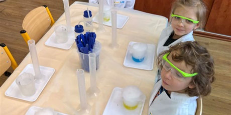Powiększ grafikę: Zajęcia chemiczne w grupie III - dzieci w białych fartuchach i okularach siedzą przy stolikach i przeprowadzają doświadczenia chemiczne