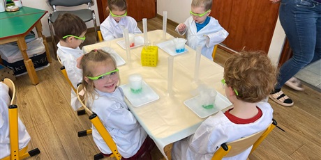 Powiększ grafikę: Zajęcia chemiczne w grupie III - dzieci w białych fartuchach i okularach siedzą przy stolikach i przeprowadzają doświadczenia chemiczne