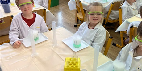 Powiększ grafikę: Zajęcia chemiczne w grupie III - dzieci w białych fartuchach i okularach siedzą przy stolikach i przeprowadzają doświadczenia chemiczne