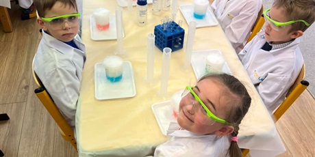 Powiększ grafikę: Zajęcia chemiczne w grupie III - dzieci w białych fartuchach i okularach siedzą przy stolikach i przeprowadzają doświadczenia chemiczne