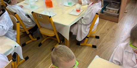 Powiększ grafikę: Zajęcia chemiczne w grupie III - dzieci w białych fartuchach i okularach siedzą przy stolikach i przeprowadzają doświadczenia chemiczne