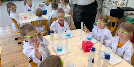 Powiększ grafikę: Zajęcia chemiczne w grupie III - dzieci w białych fartuchach i okularach siedzą przy stolikach i przeprowadzają doświadczenia chemiczne