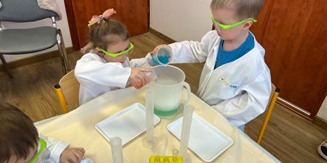 Powiększ grafikę: Zajęcia chemiczne w grupie III - dzieci w białych fartuchach i okularach siedzą przy stolikach i przeprowadzają doświadczenia chemiczne