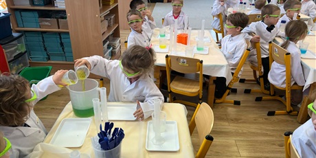 Powiększ grafikę: Zajęcia chemiczne w grupie III - dzieci w białych fartuchach i okularach siedzą przy stolikach i przeprowadzają doświadczenia chemiczne