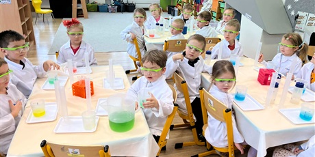 Powiększ grafikę: Zajęcia chemiczne w grupie III - dzieci w białych fartuchach i okularach siedzą przy stolikach i przeprowadzają doświadczenia chemiczne