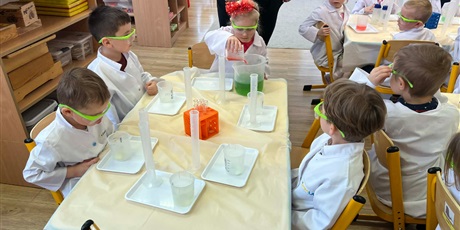 Powiększ grafikę: Zajęcia chemiczne w grupie III - dzieci w białych fartuchach i okularach siedzą przy stolikach i przeprowadzają doświadczenia chemiczne