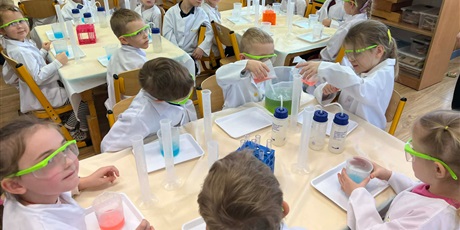 Powiększ grafikę: Zajęcia chemiczne w grupie III - dzieci w białych fartuchach i okularach siedzą przy stolikach i przeprowadzają doświadczenia chemiczne