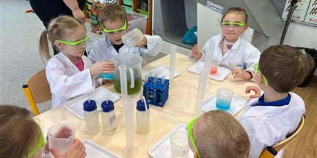 Powiększ grafikę: Zajęcia chemiczne w grupie III - dzieci w białych fartuchach i okularach siedzą przy stolikach i przeprowadzają doświadczenia chemiczne