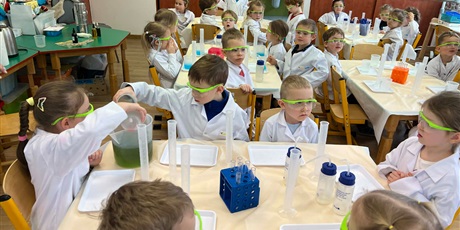 Powiększ grafikę: Zajęcia chemiczne w grupie III - dzieci w białych fartuchach i okularach siedzą przy stolikach i przeprowadzają doświadczenia chemiczne