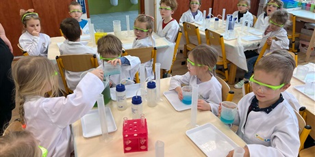 Powiększ grafikę: Zajęcia chemiczne w grupie III - dzieci w białych fartuchach i okularach siedzą przy stolikach i przeprowadzają doświadczenia chemiczne