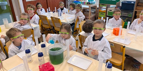 Powiększ grafikę: Zajęcia chemiczne w grupie III - dzieci w białych fartuchach i okularach siedzą przy stolikach i przeprowadzają doświadczenia chemiczne