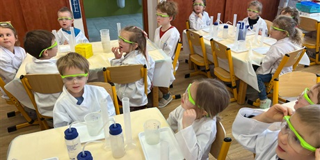 Powiększ grafikę: Zajęcia chemiczne w grupie III - dzieci w białych fartuchach i okularach siedzą przy stolikach i przeprowadzają doświadczenia chemiczne