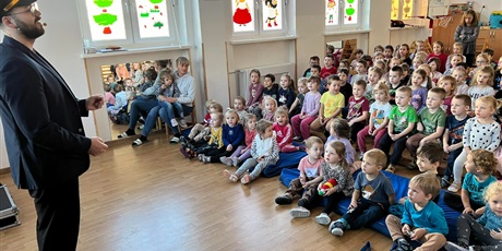 Powiększ grafikę: Dzieci z całego przedszkola podczas Koncertu na sali gimnastycznej siedzą na materacach i ławeczkach. Z lewej strony pan aktor w stroju kolejarza.