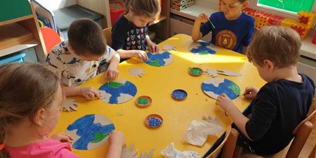 Powiększ grafikę: Przy stoliku siedzą dzieci z grupy II i malują farbami Ziemię