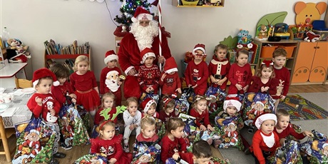 Powiększ grafikę: Na zdjęciu Krasnoludki z Mikołajem. Dzieci ubrane na czerwono w czapeczkach mikołajkowych trzymają siatki z prezentami. 