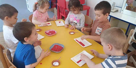Powiększ grafikę: Na zdjęciu dzieci siedzą przy stoliku i wyklejają swoje flagi białymi i czerwonymi kulkami papieru