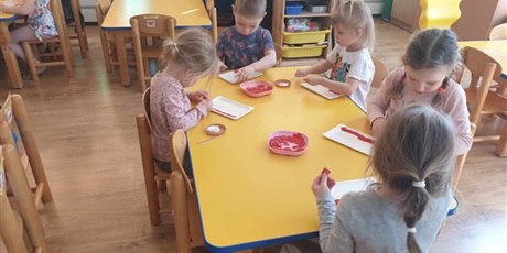 Powiększ grafikę: Na zdjęciu dzieci siedzą przy stoliku i wyklejają swoje flagi białymi i czerwonymi kulkami papieru