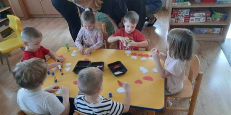 Powiększ grafikę: Na zdjęciu dzieci z grupy II z kolegami i koleżankami z Przedszkola nr 40 siedzą przy stolikach i wykonują kotyliony z białych i czerwonych serduszek.