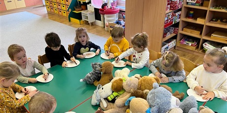 Powiększ grafikę: Dzieci siedzą przy stoliku i wykonują samodzielnie misie z talerzyków jednorazowych. Na środku stołów leżą misie, któe dziś przyniosły dzieci do przedszkola.