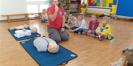 Powiększ grafikę: Zajęcia edukacyjne z udzielania pierwszej pomocy - grupa III - na zdjęciu dzieci siedzą na sali gimnastycznej na podłodze i uważnie słuchają pani instruktorki i uczą się jak przeprowadzić sztuczne oddychanie